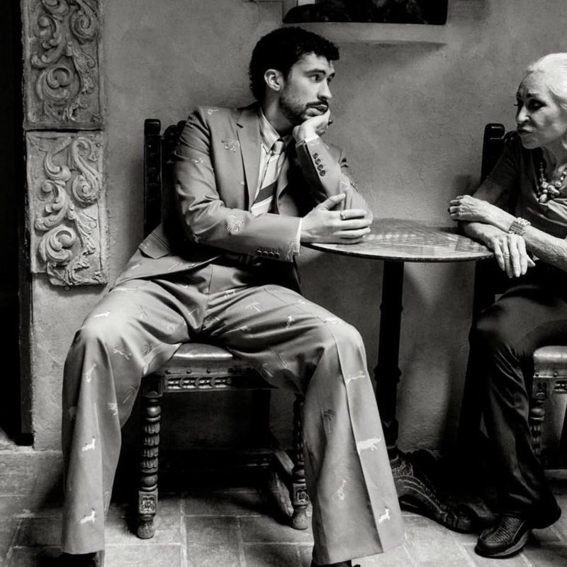 A candlelit black-and-white photograph of Bad Bunny seated with Jan D'Esopo at a small table by a carved stone relief.
