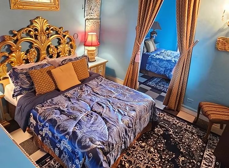 The Balcony Suite at The Gallery Inn - an ornate bedroom with a gilded headboard, blue patterned bedspread, and draped windows.