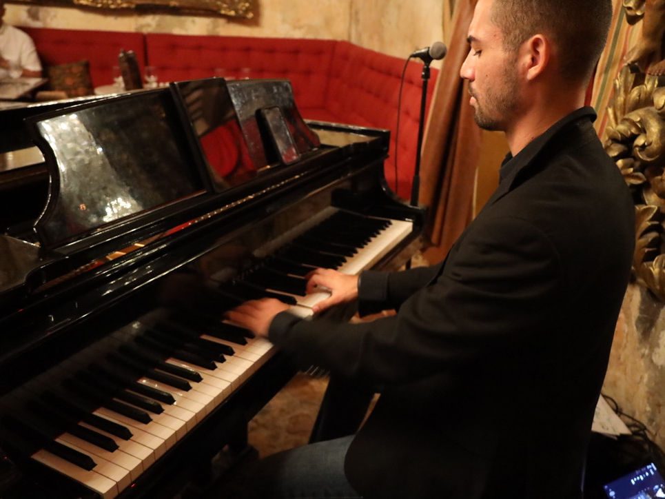 A pianist playing the grand piano at The Cannon Club, with red velvet banquettes behind.