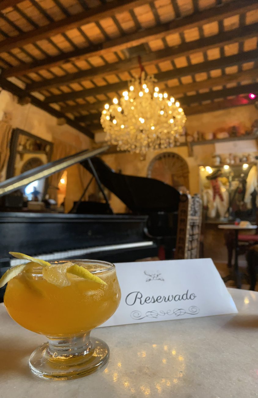 A candlelit cocktail on a marble table at The Cannon Club, with a 'Reservado' card, a black grand piano, a crystal chandelier, and a wood-beam ceiling beyond.