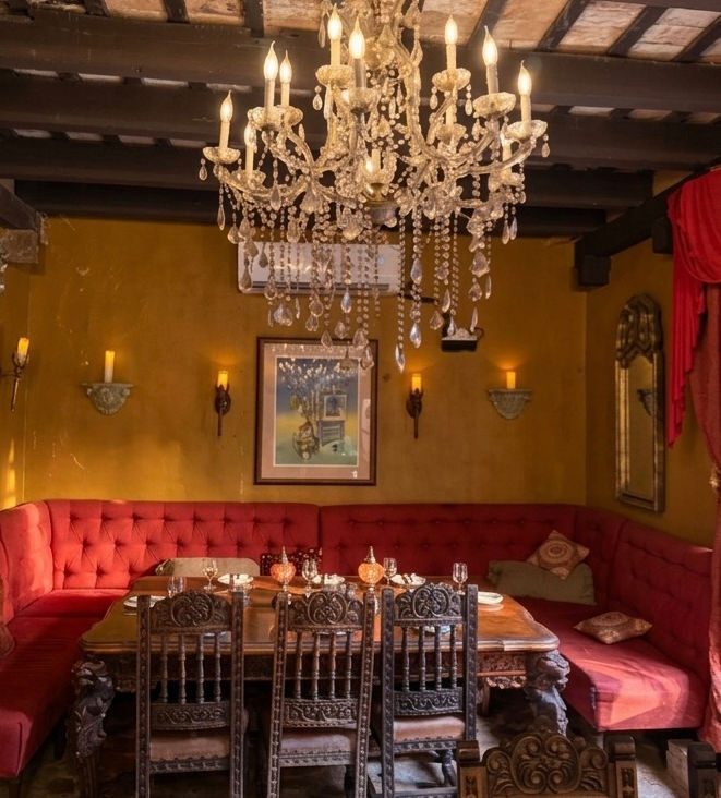 A candlelit dining nook at The Cannon Club - a crystal chandelier over a red velvet banquette, carved dark-wood chairs, and a table set with wine glasses.