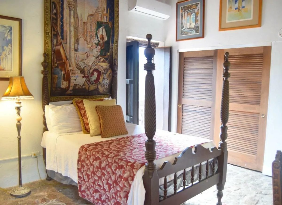 A Colonial Room at The Gallery Inn - a carved four-poster bed, a woven tapestry on the wall, louvered wood shutters, and warm plaster walls.