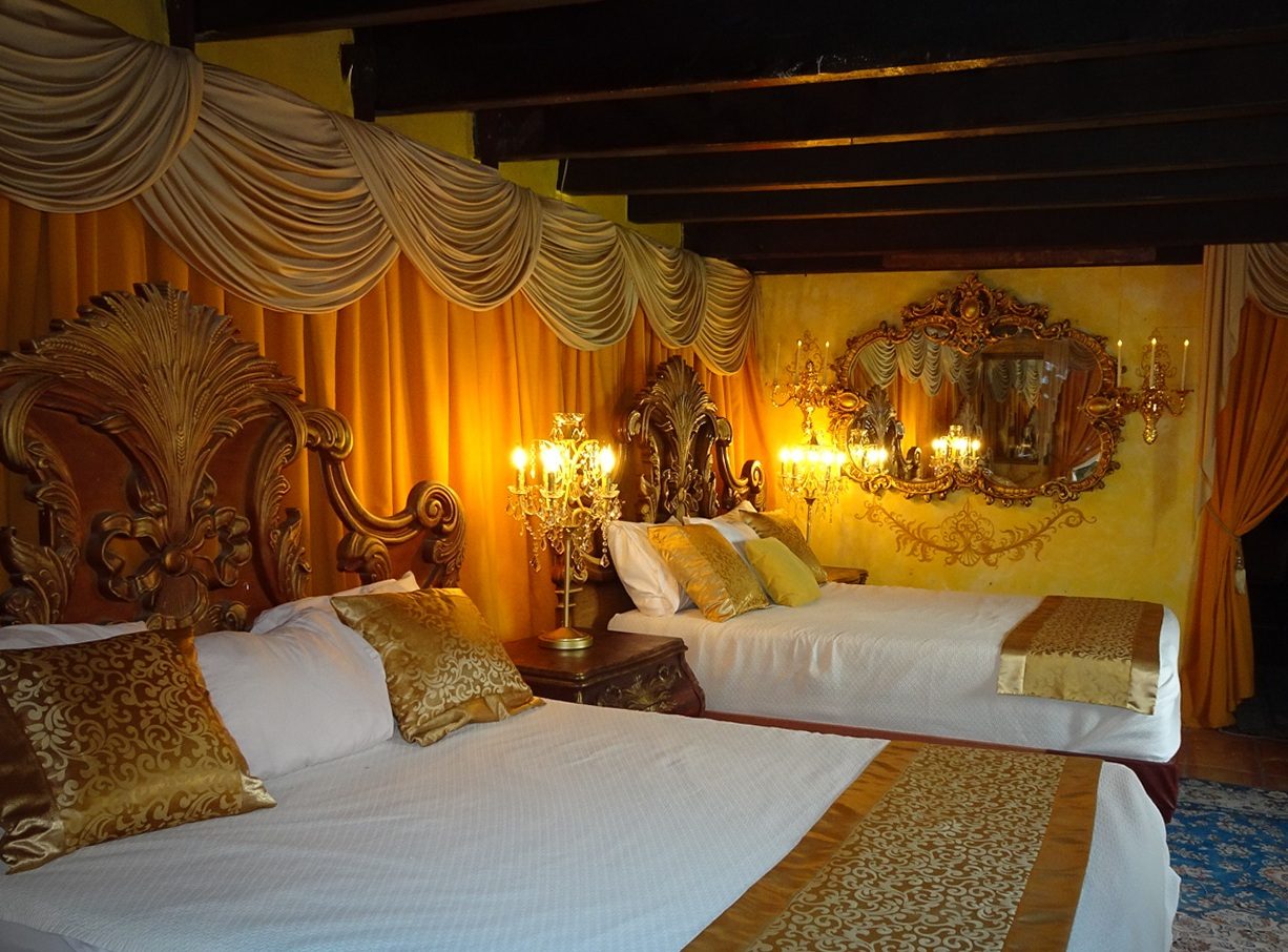 The Double Suite at The Gallery Inn - two queen beds with carved gilded headboards under gold drapery, candlelight, and an ornate mirror on the ochre wall.