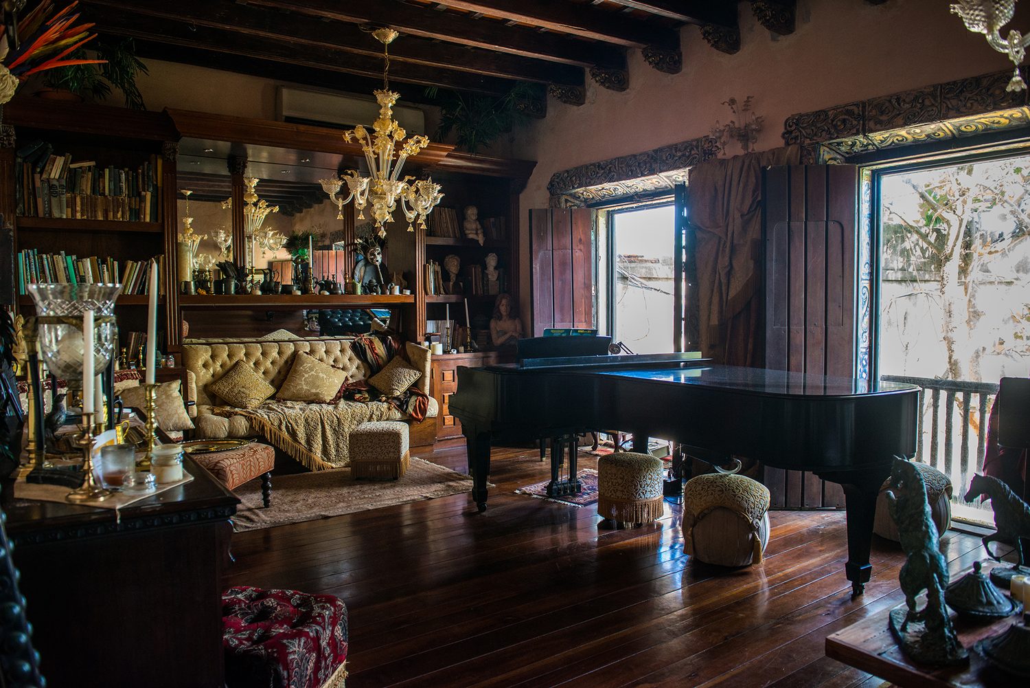 The Music Room at The Gallery Inn - a black grand piano on a dark-wood floor, with a tufted sofa, chandelier, and windows to the courtyard.