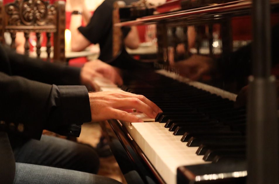 A close-up of a pianist's hands on the keys by candlelight, with a carved antique chair and warm red light behind, during a live performance at The Gallery Inn.