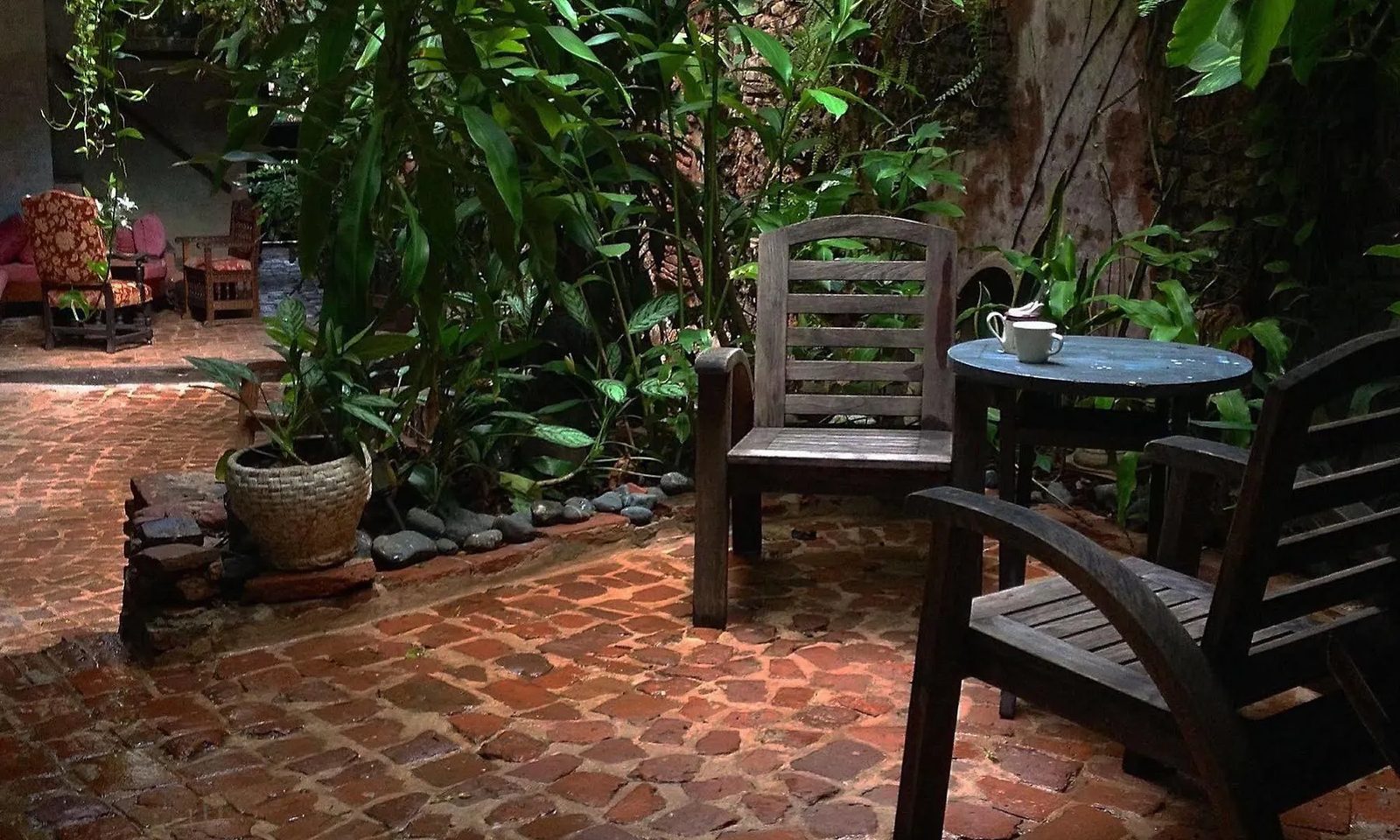 A hidden brick patio with an antique wooden chair, a small table set with a teapot, and lush tropical plants.