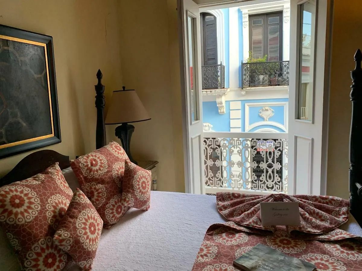 A guest room at The Gallery Inn with a patterned terracotta bedspread and an open wrought-iron balcony framing a blue Spanish-colonial façade across the Old San Juan street.