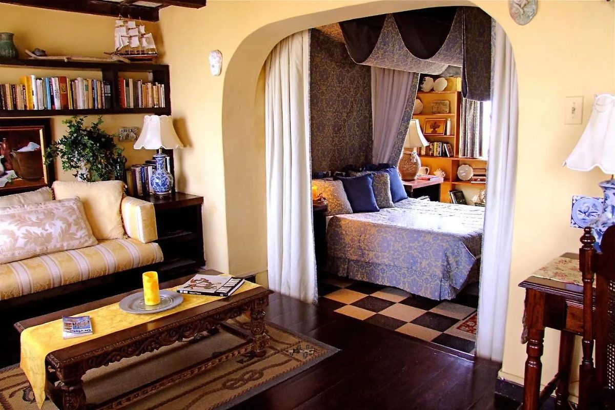 A suite parlor at The Gallery Inn - a sofa and bookshelves beside an archway to a canopied bed alcove, warm with lamplight and dark wood.