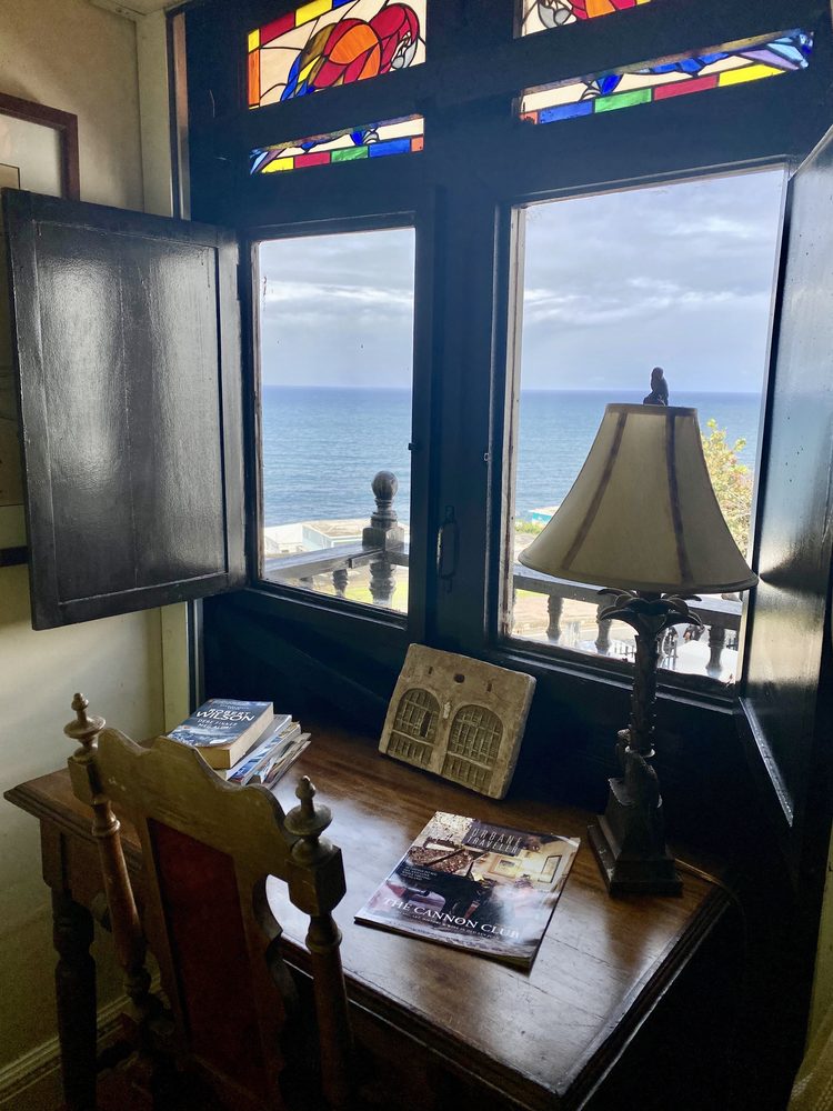 A writing desk by a shuttered window at The Gallery Inn - a stained-glass transom above and the open Atlantic beyond.