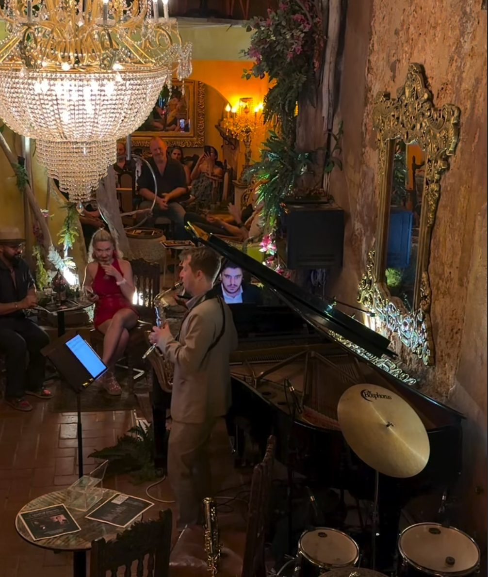 A live combo in Studio Hall at The Gallery Inn - a singer in red, a saxophonist, and a grand piano under a crystal chandelier.