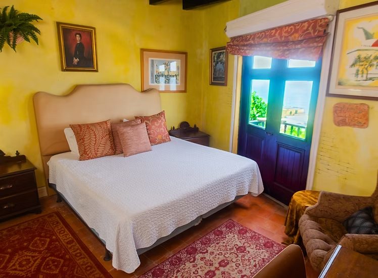 A Vista Room at The Gallery Inn - ochre walls, a large bed, wood-beam ceiling, terracotta floor, and green shutter doors to a balcony.